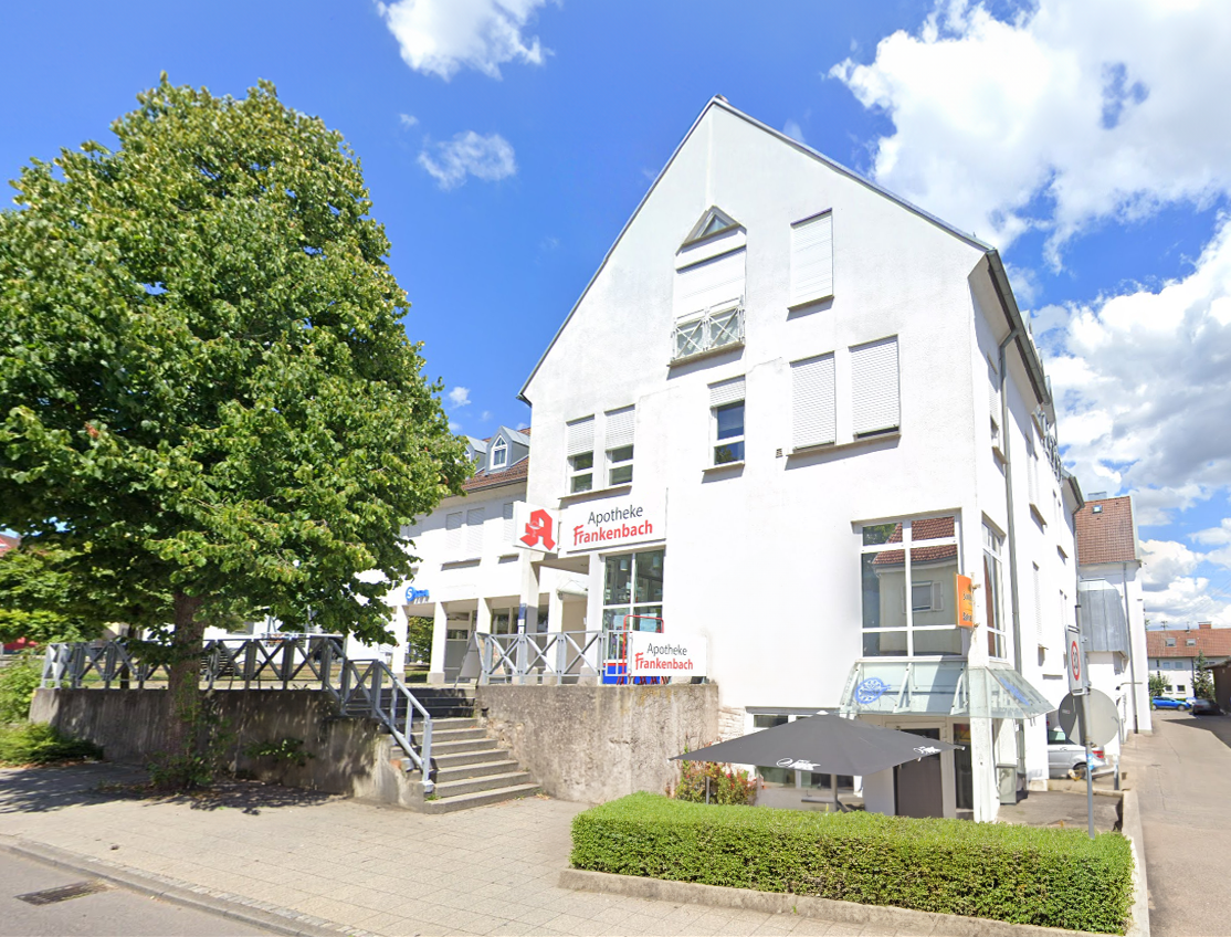 Außenansicht der Apotheke Frankenbach in Heilbronn – weißes Gebäude mit rotem Apotheken-Logo und Schriftzug, umgeben von Grünflächen an einem sonnigen Tag.