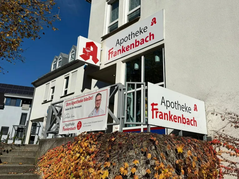 Außenansicht der Apotheke Frankenbach an einem sonnigen Tag. Das weiße Gebäude zeigt mehrere rote Logos und Schriftzüge mit dem Namen 'Apotheke Frankenbach'. Ein großes, stilisiertes rotes 'A' (das deutsche Apotheken-Logo) ragt als Schild aus der Fassade. Am Geländer hängt ein weißes Werbebanner mit dem Porträt eines Mannes in Brille und Laborkittel sowie der Aufschrift 'Ihre Apotheke bleibt! Danke für Ihr Vertrauen.' Im Vordergrund ist eine Steinmauer zu sehen, die dicht mit herbstlich gefärbtem, gelb-orangefarbenem Weinlaub bewachsen ist. Links führen Steinstufen nach oben.